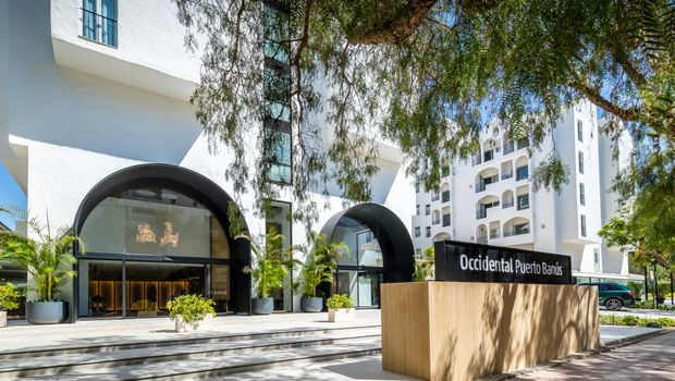Eingangsbereich des Occidental Puerto Banús Hotels mit modernem Design, großen Glasbögen, gepflegter Begrünung und Hotelnamen auf einem stilvollen Schild.