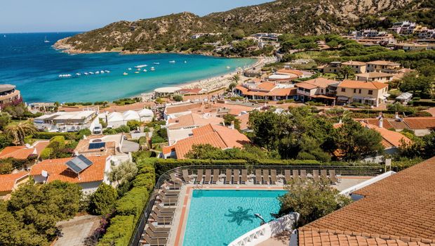 Panoramablick auf eine traumhafte Bucht mit türkisblauem Meer, feinem Sandstrand und malerischem Küstenort – im Vordergrund ein Hotelpool mit Liegestühlen.
