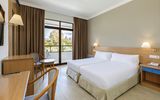 Helles Hotelzimmer mit zwei Einzelbetten, Schreibtisch, Sitzbereich und Zugang zu einem Balkon mit Blick auf eine grüne Gartenlandschaft.