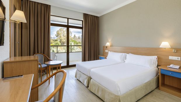 Helles Hotelzimmer mit zwei Einzelbetten, Schreibtisch, Sitzbereich und Zugang zu einem Balkon mit Blick auf eine grüne Gartenlandschaft.