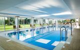 Ein moderner, überdachter Innenpool mit großen Glasfronten und Blick auf eine gepflegte Gartenanlage. Rund um den Pool stehen einzelne Stühle, die zum Verweilen einladen.
