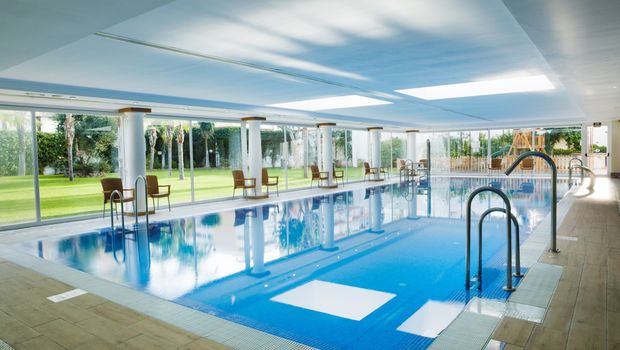Ein moderner, überdachter Innenpool mit großen Glasfronten und Blick auf eine gepflegte Gartenanlage. Rund um den Pool stehen einzelne Stühle, die zum Verweilen einladen.