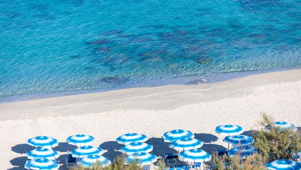 Reihe von blau-weißen Sonnenschirmen an einem feinsandigen Strand mit kristallklarem Wasser in verschiedenen Türkistönen.