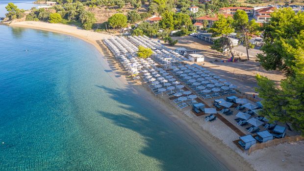 Breiter Sandstrand mit glasklarem Wasser und zahlreichen Sonnenliegen mit Schirmen, umgeben von grüner Natur und kleinen Hotelanlagen – perfekter Ort für entspannte Badetage.