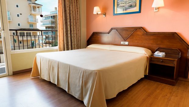Ein einfach eingerichtetes Hotelzimmer mit einem großen Doppelbett, orangener Wand, Holzfußboden und klassischem Holzmobiliar, mit Blick aus dem Fenster auf benachbarte Gebäude.