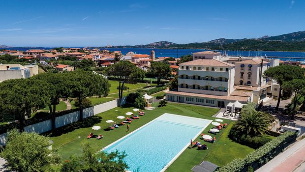 Luftaufnahme eines stilvollen Hotels mit großem Außenpool inmitten gepflegter Gartenanlagen – mit Blick auf die umliegenden Häuser, das tiefblaue Meer und die grünen Hügel Sardiniens.