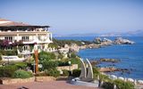 Mediterranes Hotelgebäude direkt an der felsigen Küste mit traumhaftem Blick auf das türkisfarbene Meer und vorgelagerte Inseln – üppige Blumen und grüne Gartenanlagen im Vordergrund.