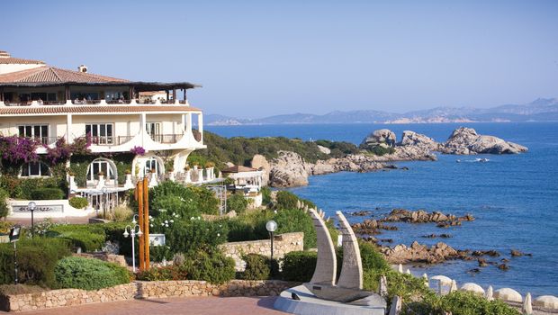 Mediterranes Hotelgebäude direkt an der felsigen Küste mit traumhaftem Blick auf das türkisfarbene Meer und vorgelagerte Inseln – üppige Blumen und grüne Gartenanlagen im Vordergrund.