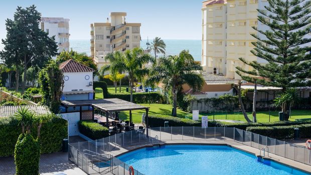 Blick auf einen runden Hotelpool mit Sicherheitszaun, daneben eine überdachte Sitzterrasse, üppige grüne Gartenanlagen mit Palmen sowie mehrstöckige Gebäude im Hintergrund, dahinter das Meer.