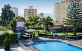 Blick auf einen runden Hotelpool mit Sicherheitszaun, daneben eine überdachte Sitzterrasse, üppige grüne Gartenanlagen mit Palmen sowie mehrstöckige Gebäude im Hintergrund, dahinter das Meer.