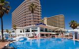 Modernes Hotelgebäude mit mehreren Etagen und geschwungenen Balkonen, direkt an einem großen Außenpool mit Liegestühlen und Sonnenschirmen, unter strahlend blauem Himmel – ideal für einen sonnigen Badeurlaub.