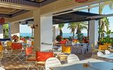 Heller, farbenfroher Außenbereich eines Restaurants mit Blick auf das Meer, umgeben von Palmen und tropischer Vegetation.