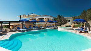 Einladender Außenpool mit türkisblauem Wasser, umgeben von blauen Sonnenschirmen und Liegestühlen vor einer mediterranen Hotelanlage unter klarem Himmel.