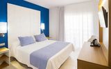 Helles Doppelzimmer mit blau-weißem Farbschema, großem Bett mit zwei blauen Zierkissen, weißen Vorhängen vor dem Balkon und einem Schreibtisch mit Wasserkocher und Flachbildfernseher.
