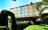 Gepflegte Gartenanlage mit geschnittenen Hecken, Palmen und einem grünen Torbogen vor einem mehrstöckigen Hotelgebäude mit Balkonen, im Hintergrund strahlend blauer Himmel.
