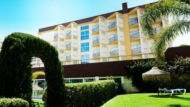 Gepflegte Gartenanlage mit geschnittenen Hecken, Palmen und einem grünen Torbogen vor einem mehrstöckigen Hotelgebäude mit Balkonen, im Hintergrund strahlend blauer Himmel.