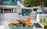 Leckeres Mittagessen mit Sandwich, Salat und Kartoffelspalten auf einem Teller, dazu ein Glas Orangensaft und ein Glas Weißwein, serviert im Freien an einem modernen Gastro-Food-Stand unter schattigen Bäumen.