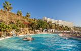 Weitläufige Poolanlage mit Wasserrutsche, Felsenlandschaft und Liegestühlen, umgeben von tropischen Palmen und blühender Vegetation, im Hintergrund ein großes Hotelgebäude an einem sonnigen Hang.