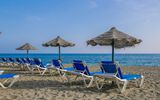 Reihe blauer Liegestühle mit Strohdach-Sonnenschirmen an einem Sandstrand, mit Blick auf das ruhige blaue Meer unter klarem Himmel.